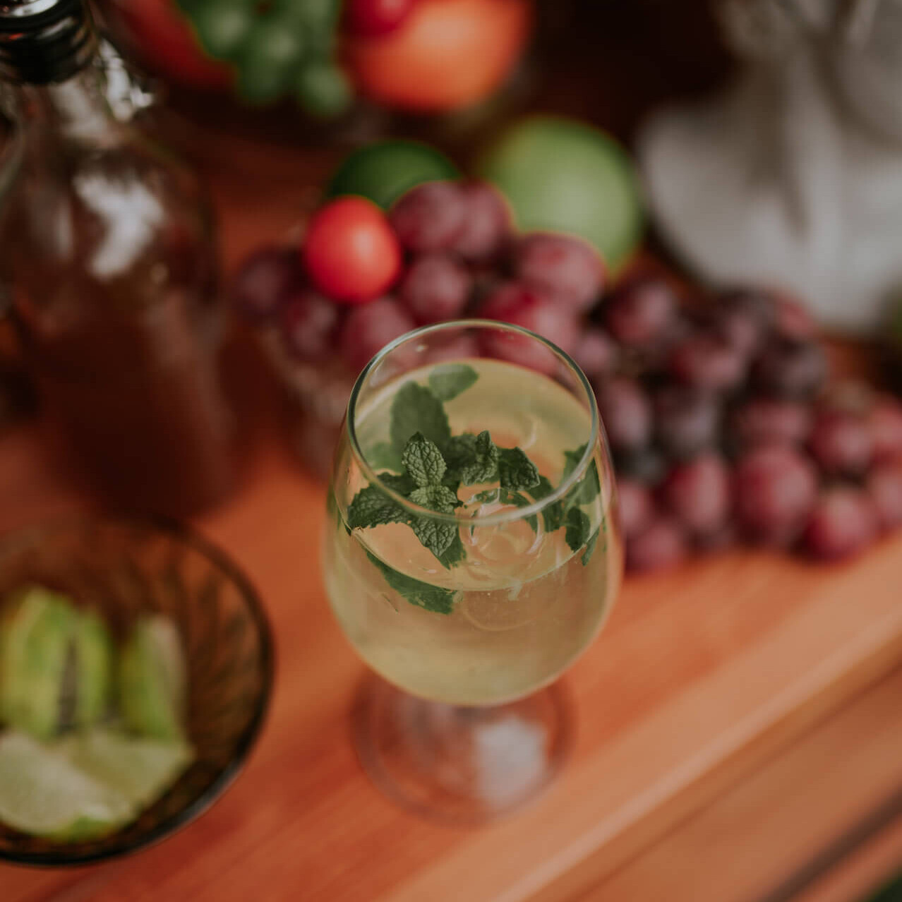 Zbliżenie na orzeźwiający, letni koktajl z miętą i lodem na drewnianym barze, w tle winogrona i świeże owoce. Oferta mobilnego drink baru i włoskich koktajli na wesela od Frizzanti.