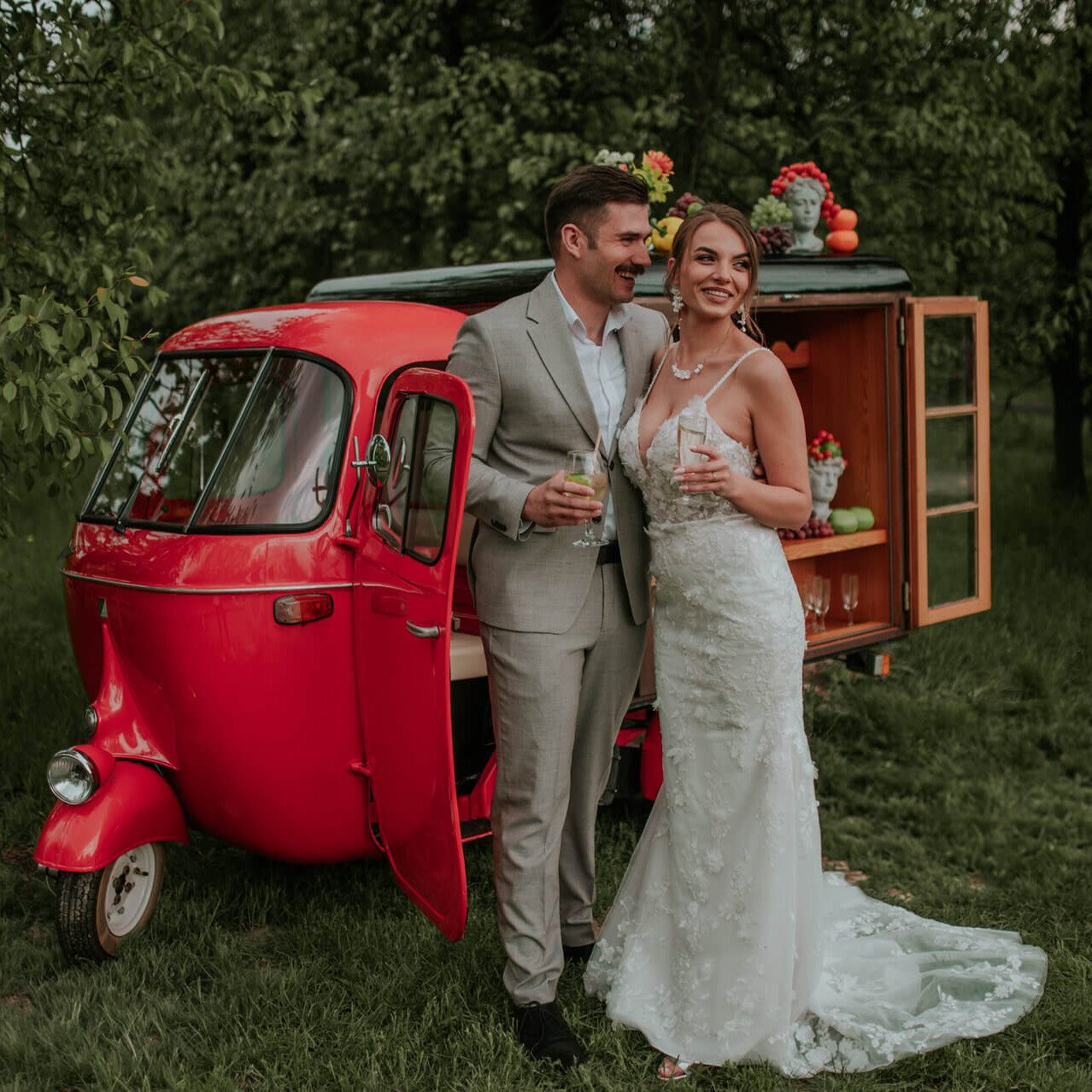 Szczęśliwi nowożeńcy wznoszący toast przy otwartym, klasycznym skuterze Piaggio Ape serwującym wino z kranu w zielonym plenerze.